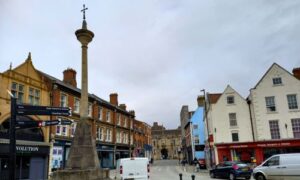 Stone cross in town square with shops and cars nearby
